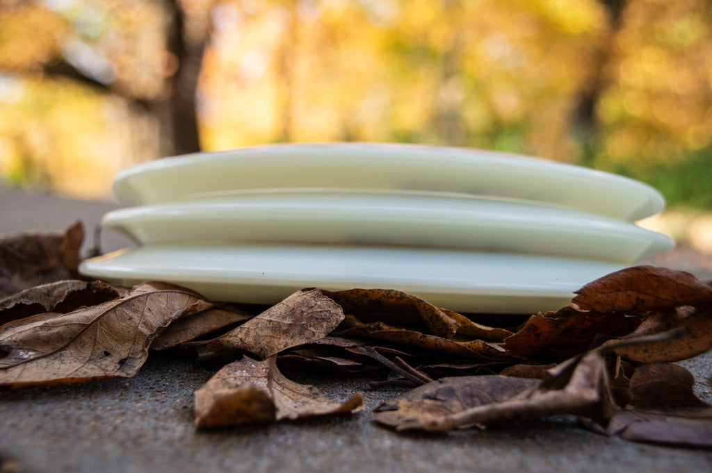 Three white discs are stacked on the ground among dry brown leaves, with a blurred background of autumn foliage.