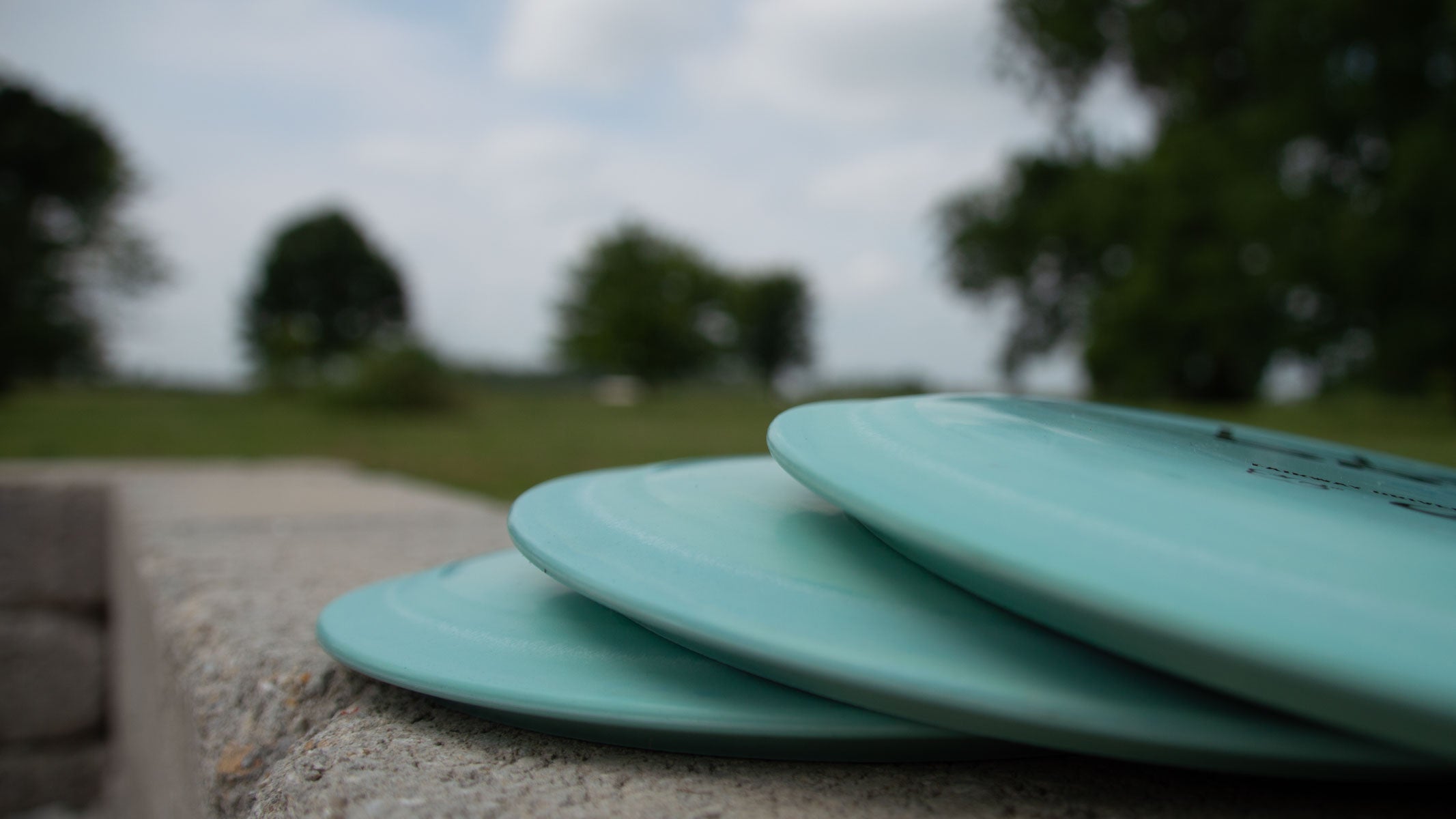 Three light blue disc golf discs are stacked on a concrete surface with a grassy park and trees blurred in the background.