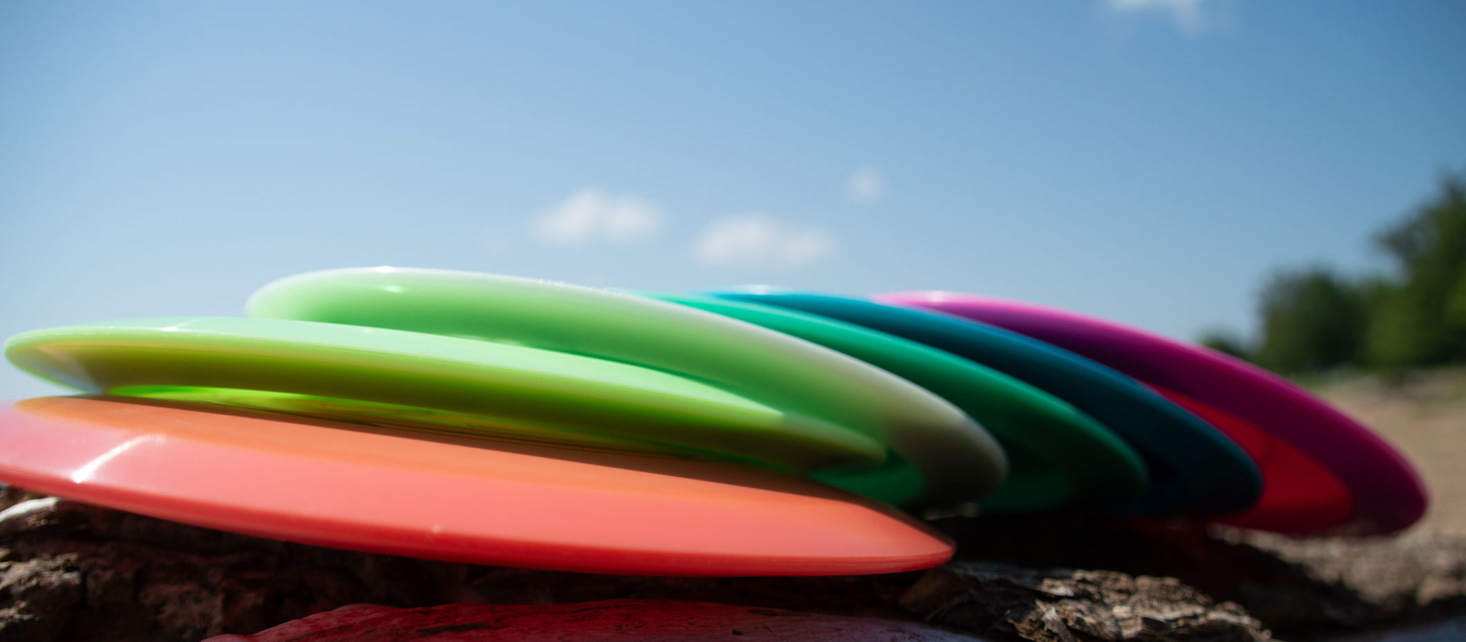 A row of colorful plastic frisbees is stacked on a rocky surface outdoors with a clear blue sky in the background.