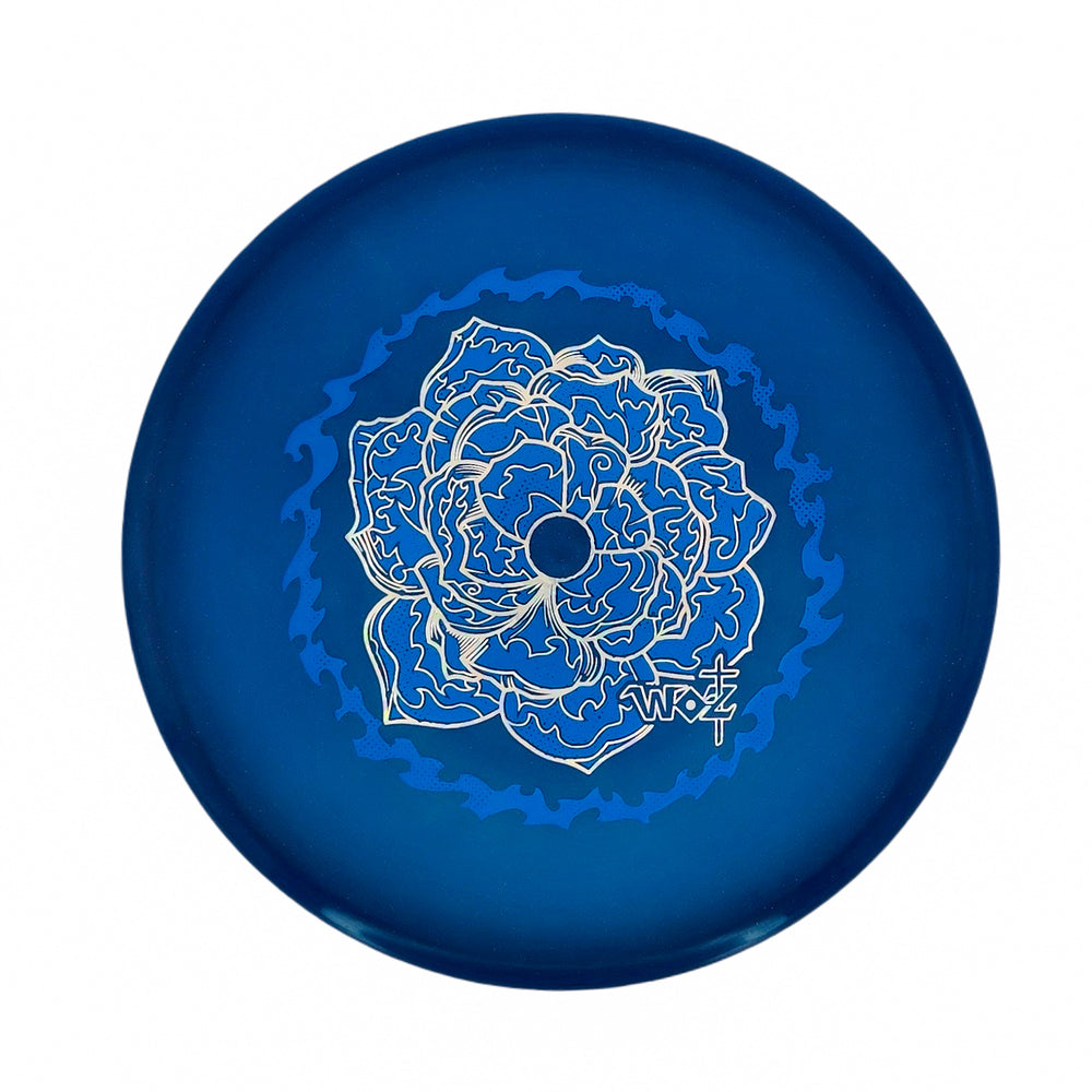 The WoznBloom Diamond Shaman is a blue disc golf disc featuring an intricate floral design in light blue and white on its surface.