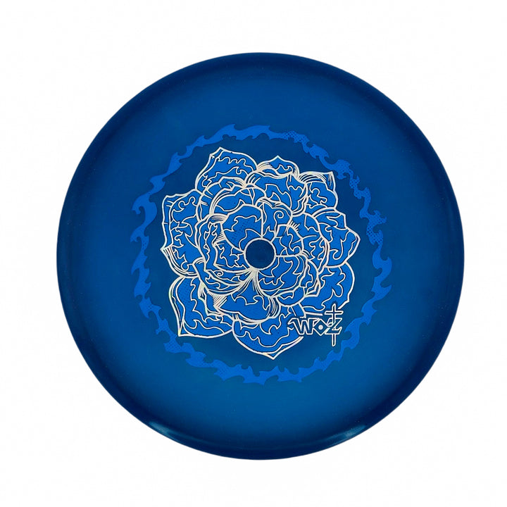 The WoznBloom Diamond Shaman is a blue disc golf disc featuring an intricate floral design in light blue and white on its surface.