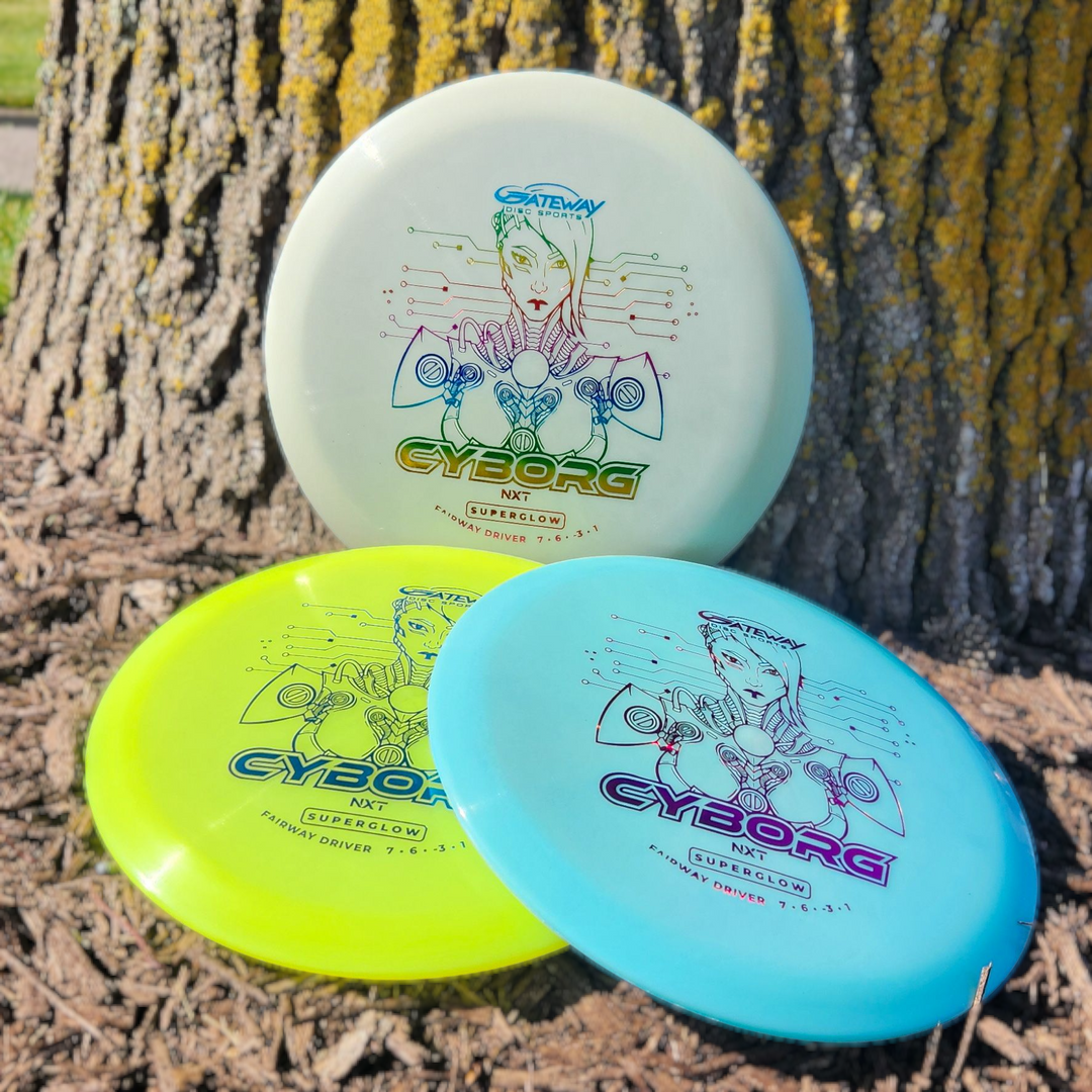 Three Cyborg - NXT Superglow discs—white, yellow, and blue—lean on a tree trunk atop mulch, each featuring a futuristic design. The set offers an understable disc and a fairway driver for versatile disc golf play.