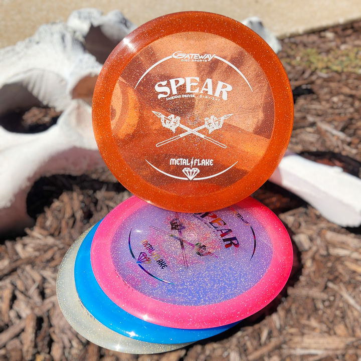 Four Gateway disc golf discs, including the orange Spear - Diamond (Metal Flake) 9-speed control driver, are stacked on mulch beside a white, weathered animal skull.