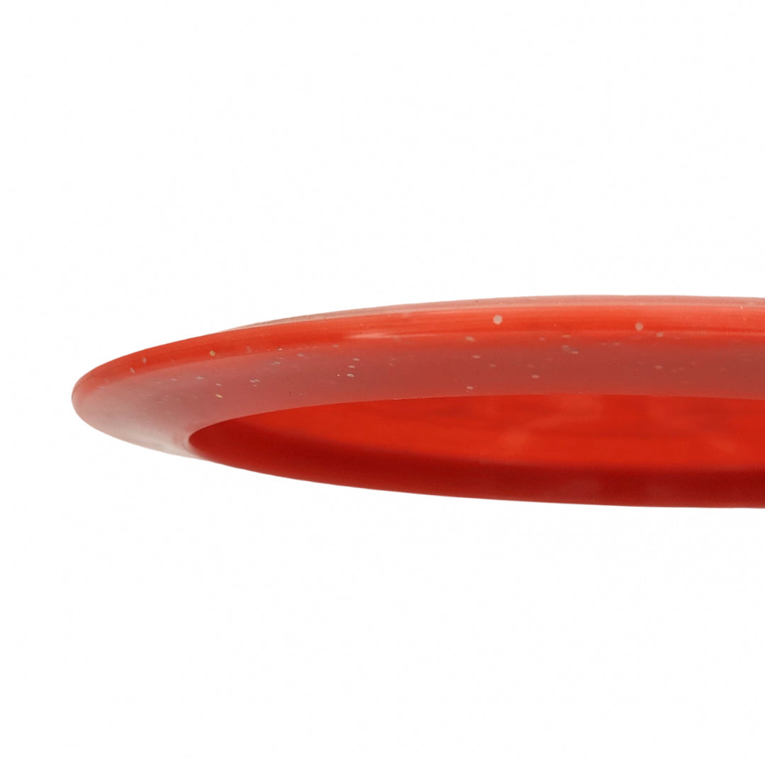 Close-up of the edge of an Ether - Diamond Metal Flake distance driver in red against a white background, perfect for max distance throws.