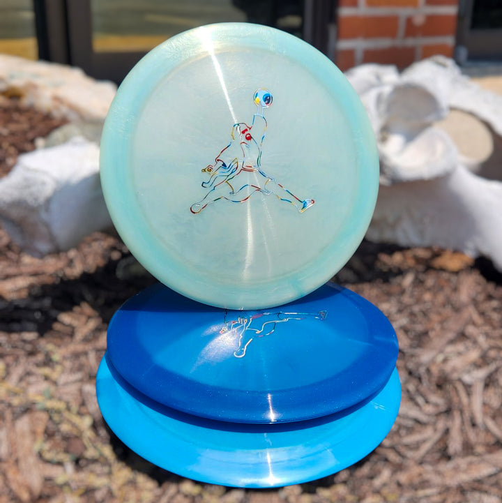 The Realm - Diamond - Air Dude, a light blue distance driver disc featuring a stylized figure logo, is shown standing atop two stacked blue discs outdoors.
