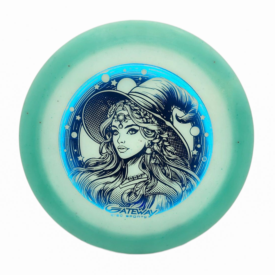 The Spell NXT Superglow - PremierJul disc features a green and white design with a blue illustration of a woman in a pointed hat with stars and moons. Crafted from premium glow plastic, it's labeled "Gateway Disc Sports.