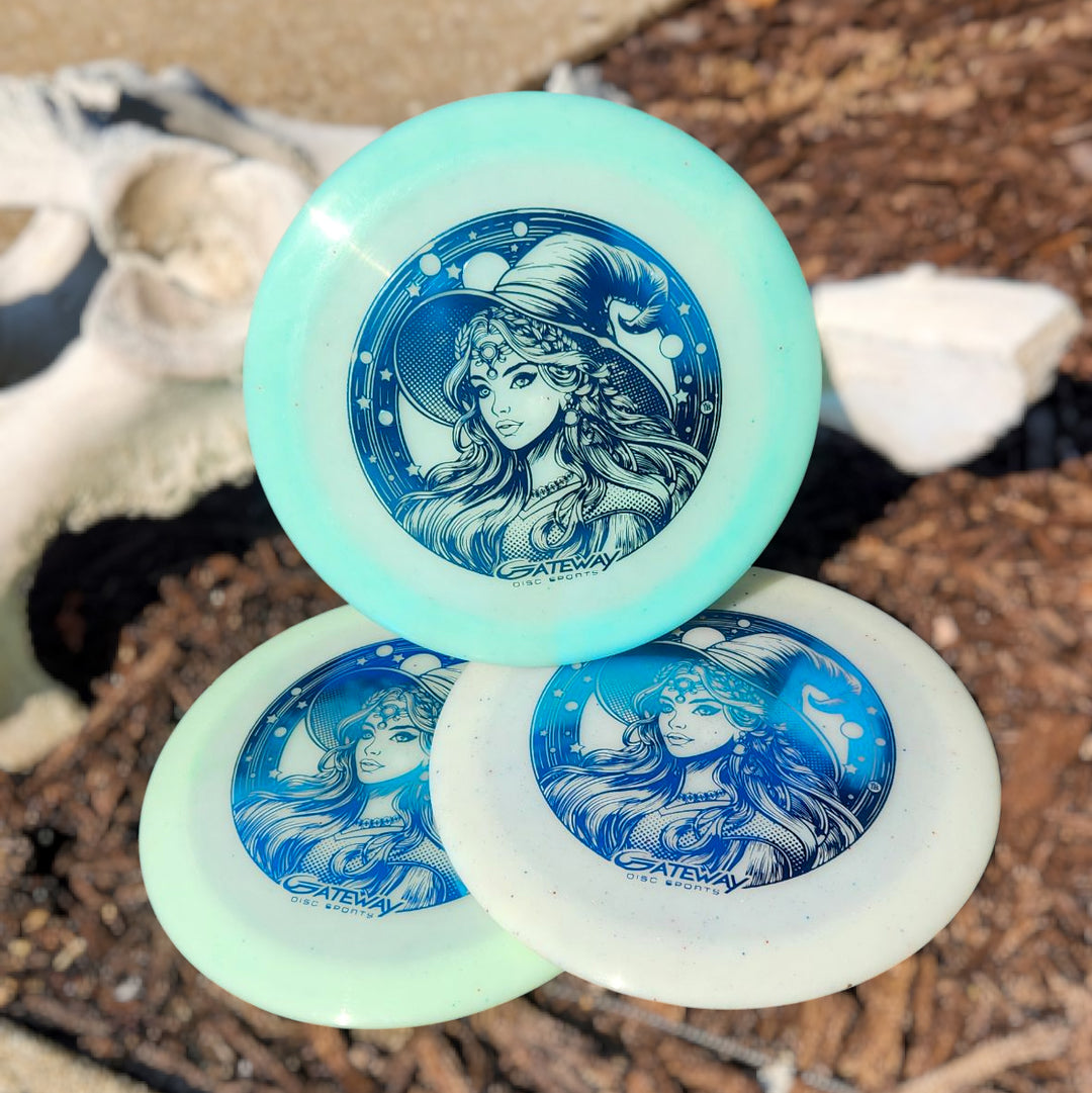 Three Spell NXT Superglow - PremierJul discs with a blue witch hat and moon design are stacked outdoors on a textured surface. Made from glow plastic, these overstable drivers shine day or night.