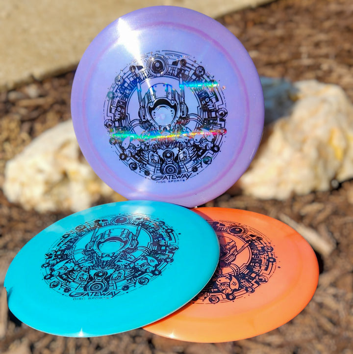 Three Spell NXT Lite - PremierJUL discs in purple, blue, and orange with black designs lie on outdoor mulch beside a large rock—perfect for power throws or as your next overstable driver.
