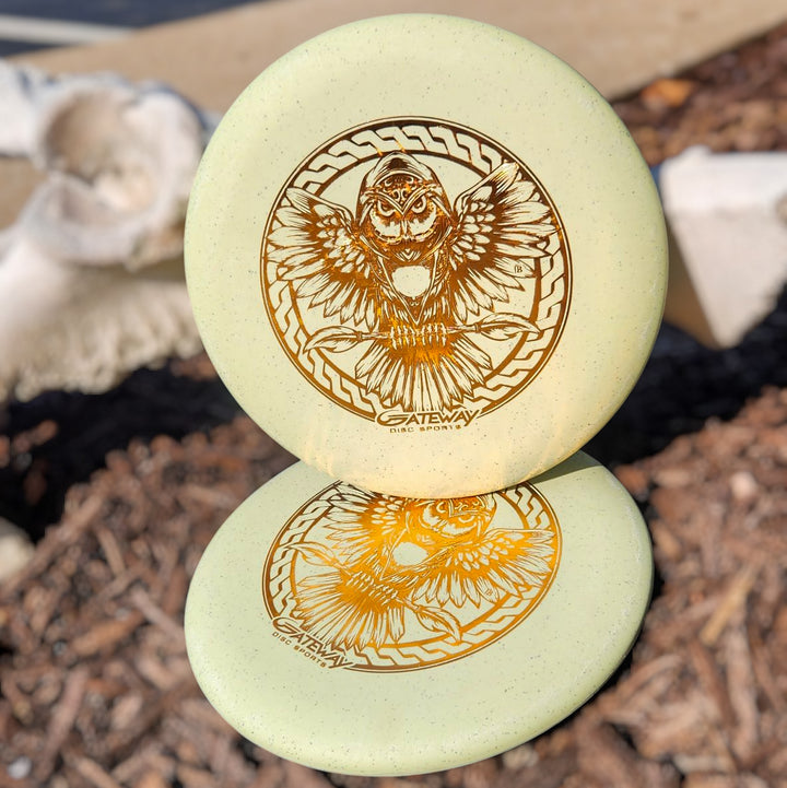 A Warlock SSS Hemp - PremierJUL disc golf putter featuring an intricate owl and Gateway logo rests outdoors, reflected on a lower disc. This light-colored, beadless putter is made from organic hemp for a super soft, unique feel.