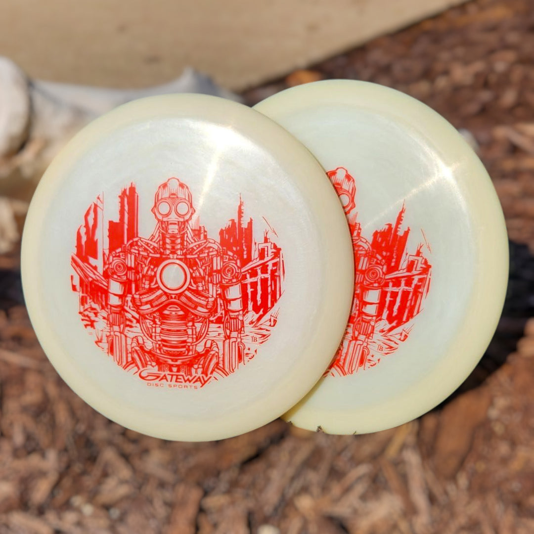 Two Cyborg Superglow - PremierJUL discs, featuring red robot and cityscape designs, lean together on wood chips outdoors—ideal for perfecting your next turnover shot.