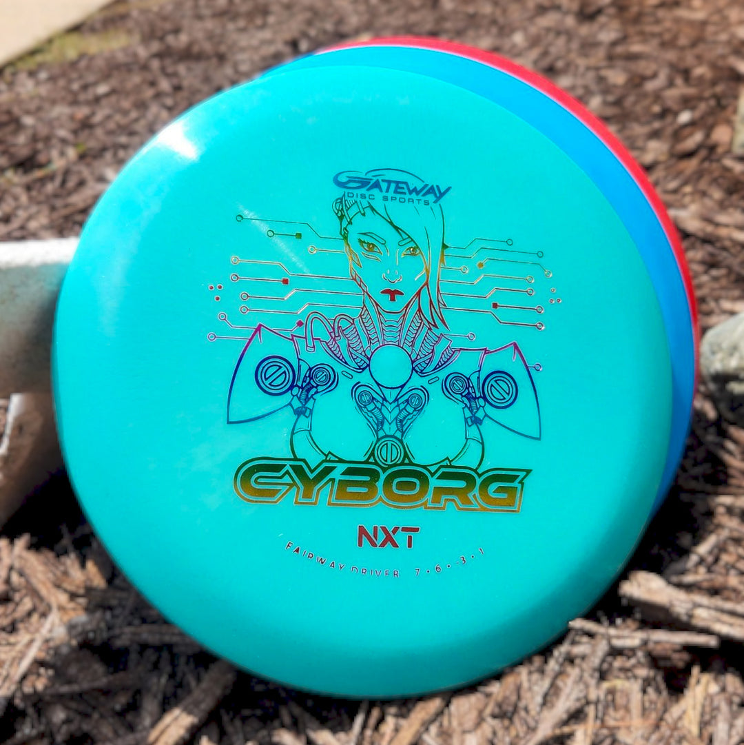 A teal "Cyborg - NXT" disc golf fairway driver featuring a futuristic robot design, resting on mulch beside two other discs.
