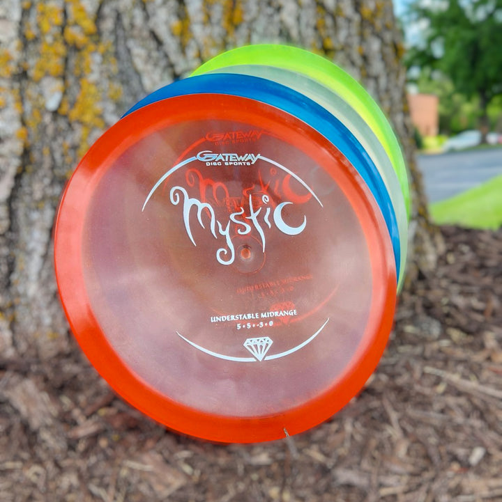 The Mystic - Diamond disc, with impressive glide and a straight flight, is stacked with two others against a tree trunk outdoors—perfect for controllable mid-range throws through greenery and mulch.