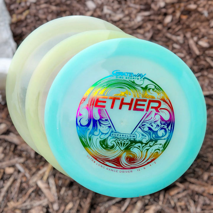 Three Gateway disc golf discs stacked on brown mulch, with the top disc labeled "Ether - Diamond Superglow"—a vibrant distance driver crafted for maximum distance shots.