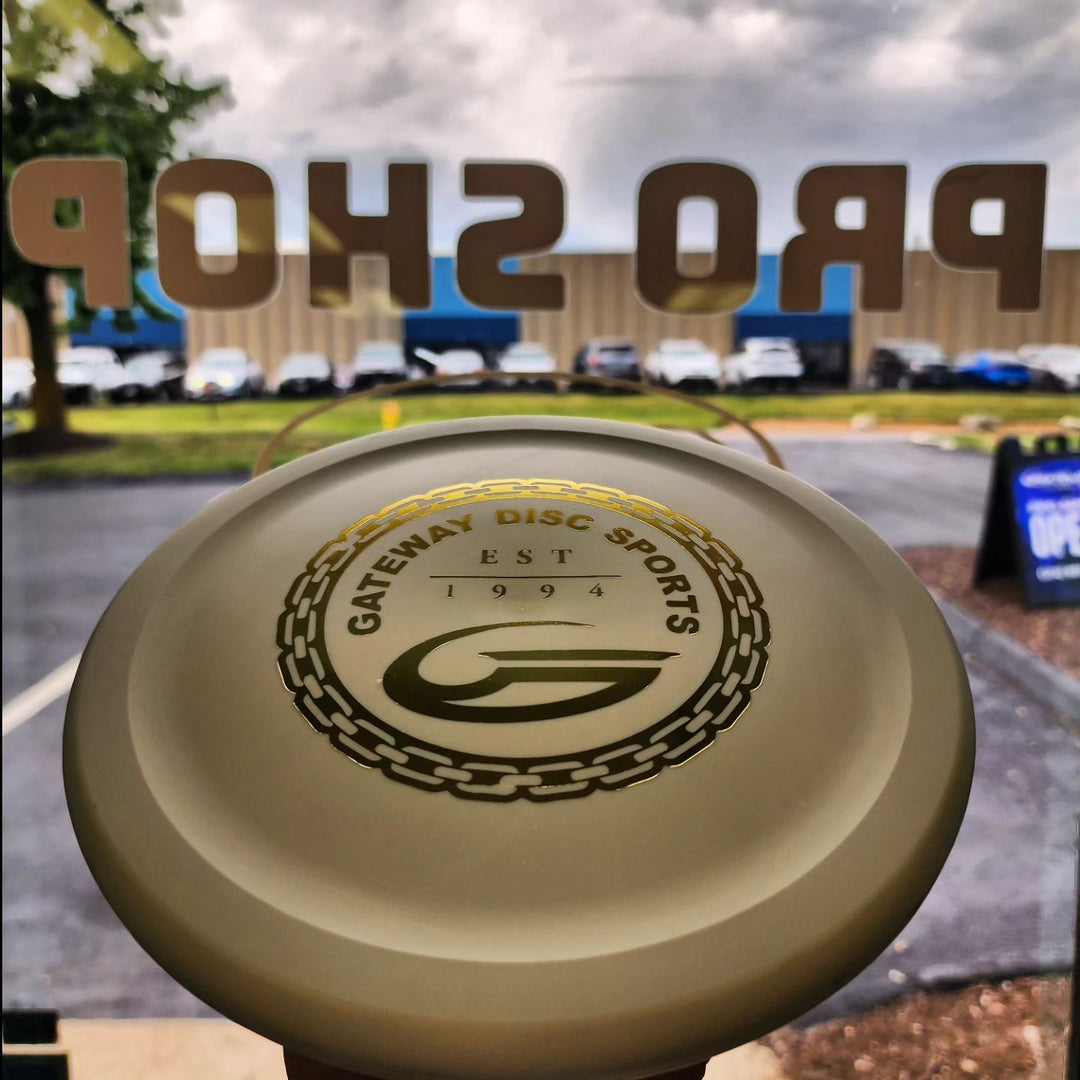 A Warspear - Prototype Suregrip putt and approach disc is held up in front of a glass door with "PRO SHOP" lettering, overlooking a parking lot and cloudy sky.
