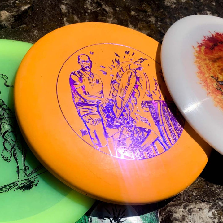 Three disc golf discs are shown, with the center orange Black Zombie Chainsaw — SS putter from BZ Sports featuring a purple illustration of a person wielding a chainsaw. This stable putter stands out with its unique design and eye-catching color.