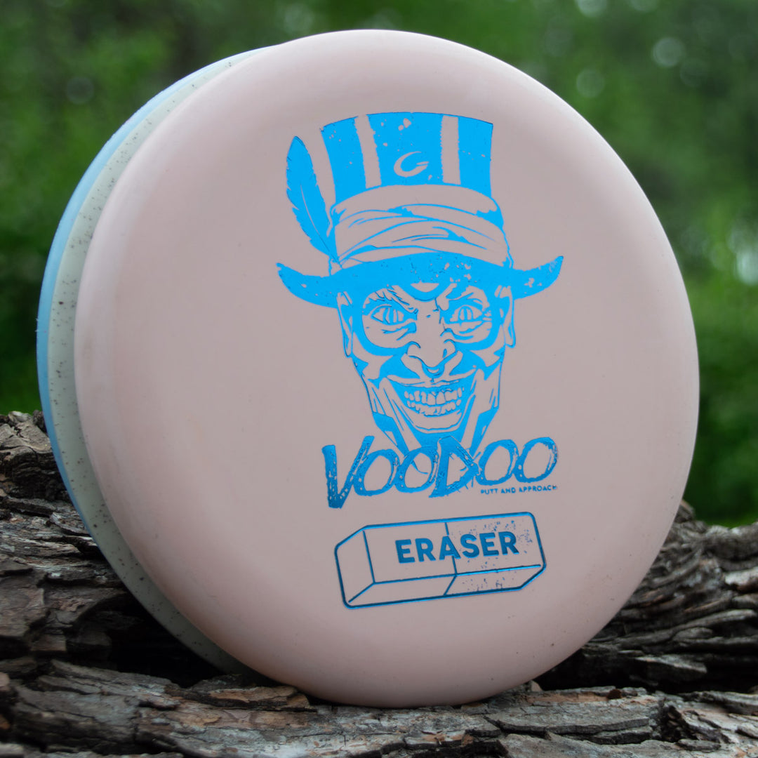 The Voodoo - Eraser Sustainability Edition is a pink putter labeled "Voodoo," with a blue grinning man graphic and "ERASER" text, shown on tree bark among greenery—a perfect lay-up disc.