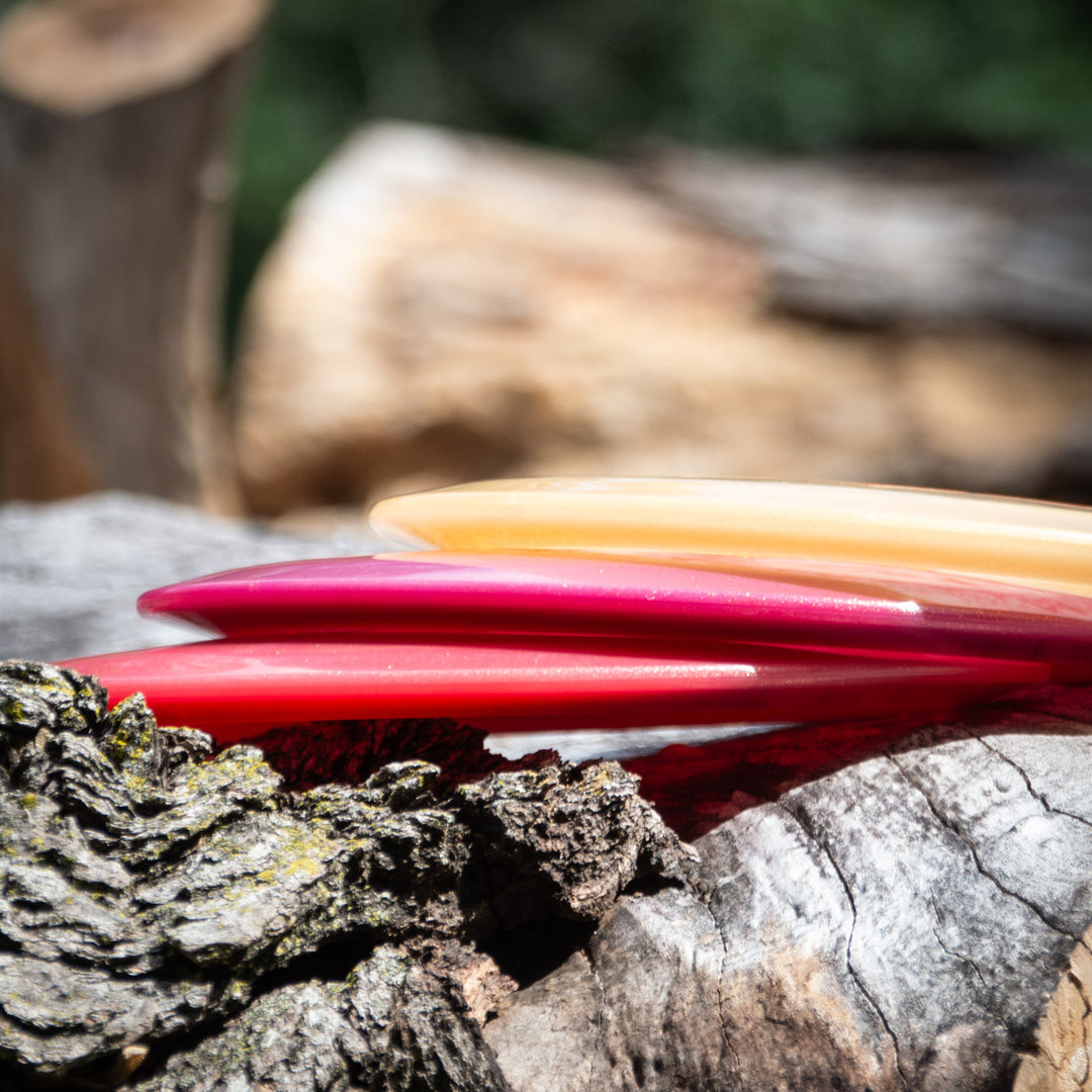 Three Lindsay Fish's Assassin - Diamond Metal Flake discs, one orange and two pink, rest on rough tree bark with a blurred natural background—perfect for your next fairway drives.