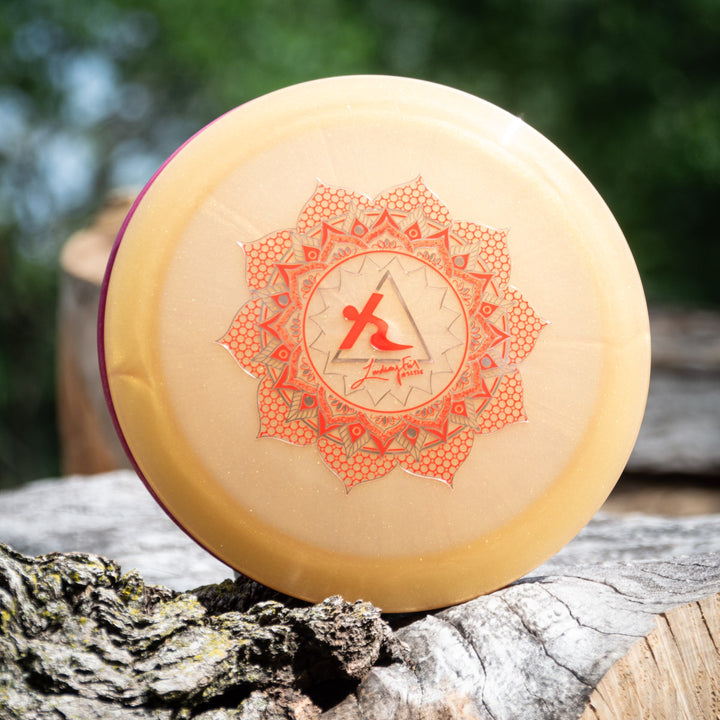 The Lindsay Fish's Assassin - Diamond Metal Flake is a beige disc golf distance driver with a red geometric mandala and signature logo, pictured resting on tree bark outdoors.