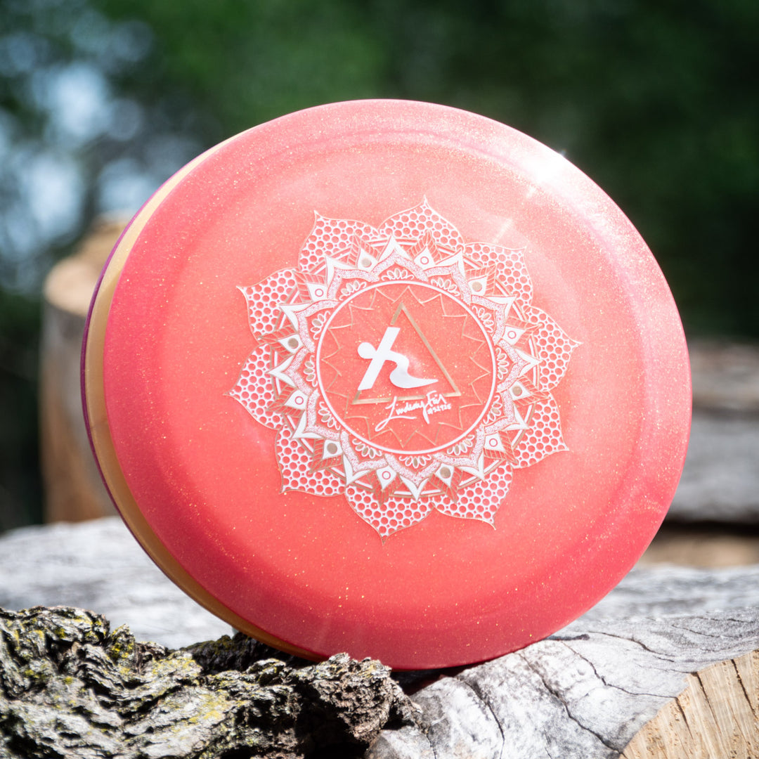 A Lindsay Fish's Assassin - Diamond Metal Flake disc golf distance driver, pink with a white geometric design and logo, sits on a tree stump outside against a backdrop of blurred greenery.