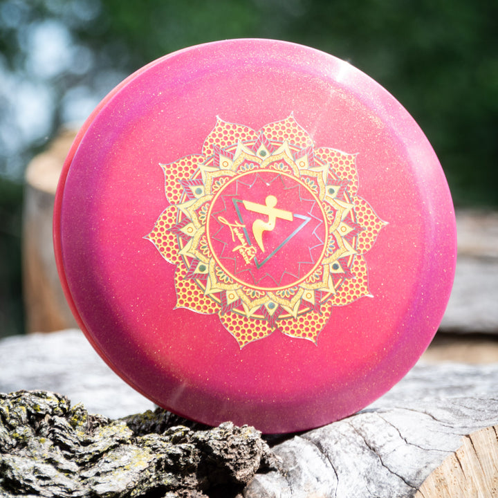 Lindsay Fish's Assassin - Diamond Metal Flake: A round, pink disc golf distance driver with a golden mandala design and a central symbol, displayed on tree bark against a blurred green background.