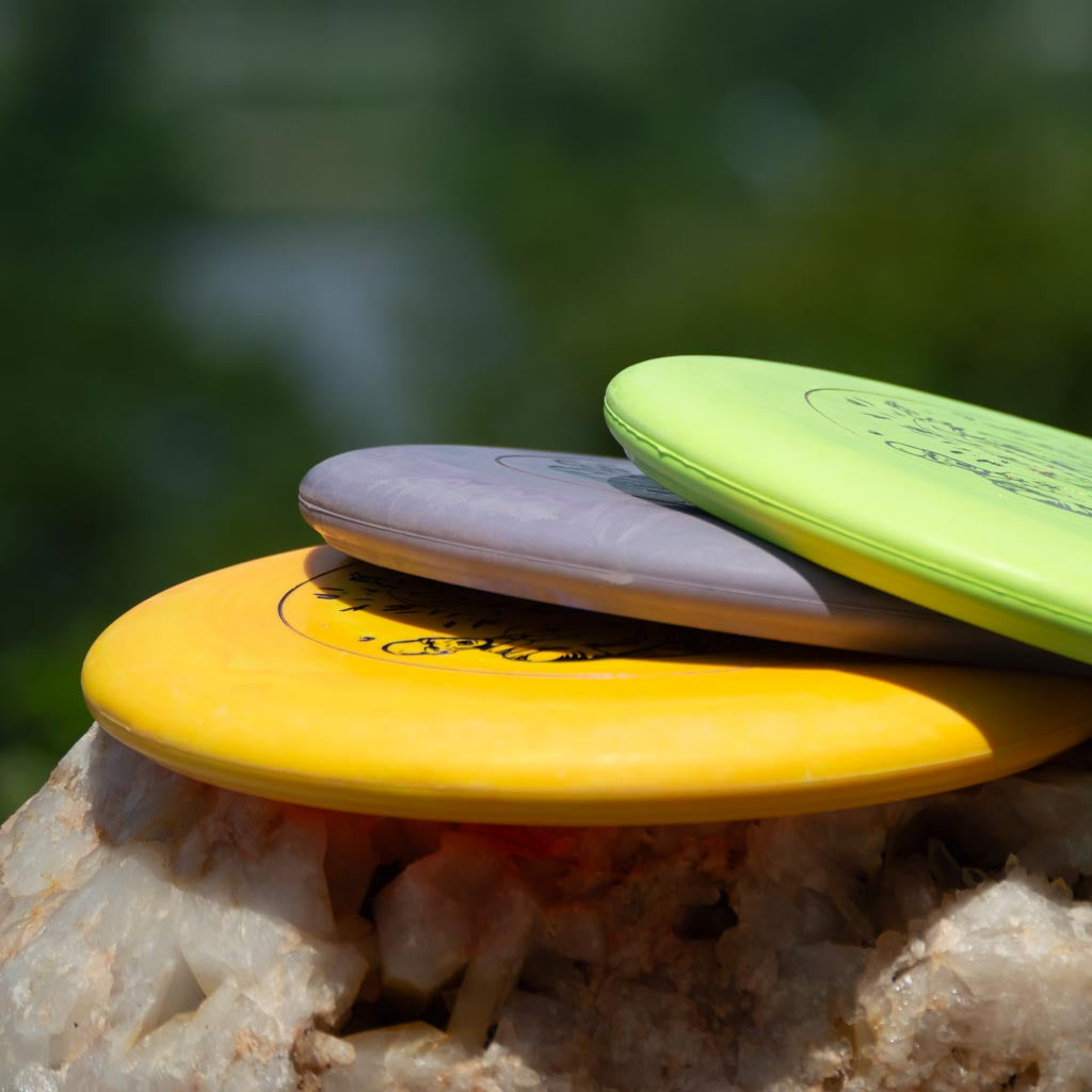 Three disc golf discs—yellow, gray Black Zombie Chainsaw — SS, and green stable putter—stacked on a rock outdoors with a blurred natural background. Perfect gear for any BZ Sports enthusiast.