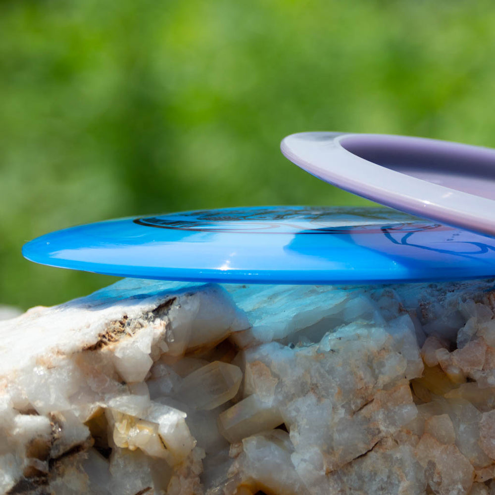 A close-up of two Black Zombie Flamethrower — Hyper Diamond discs, one blue and one purple, stacked on a rock with a green, blurred background.