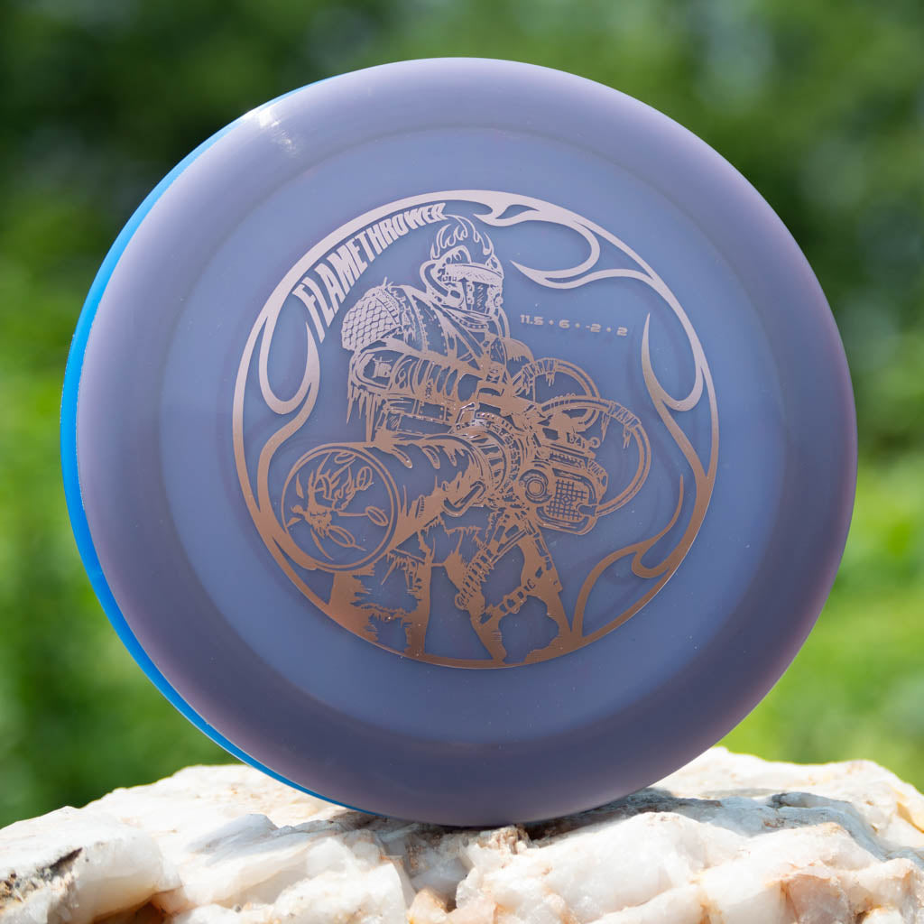 A Black Zombie Flamethrower — Hyper Diamond disc golf disc in purple and blue, engraved with a knight wielding a flamethrower, displayed on a rock outdoors.