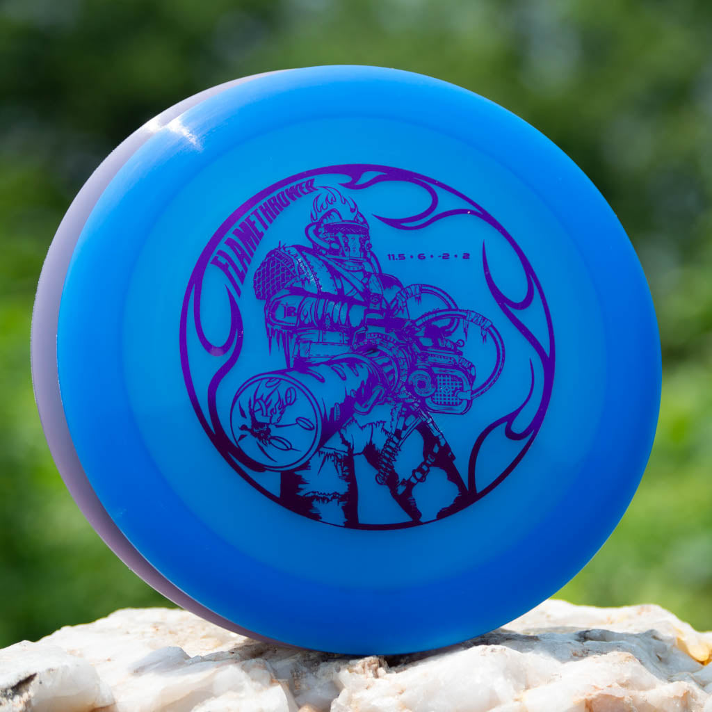 A blue Black Zombie Flamethrower — Hyper Diamond disc golf disc with a purple futuristic armored figure illustration, shown outdoors on a rock amidst greenery.