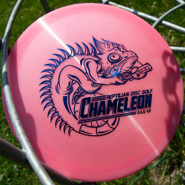 A pink Reptilian DG Chameleon — Armor fairway driver with a blue chameleon holding a disc illustration rests on a metal basket—a versatile workhorse for your disc golf game.