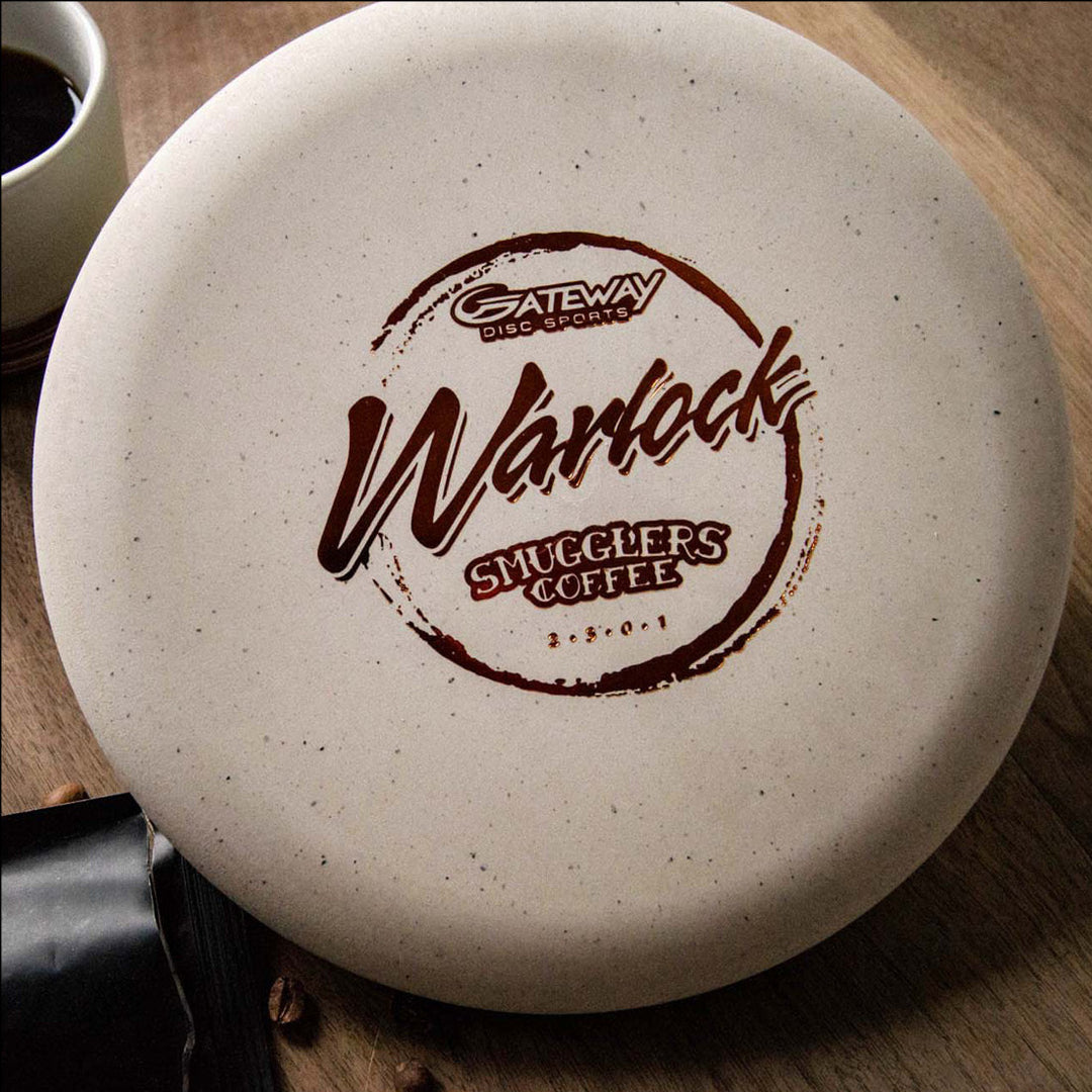 A beige Warlock - Smugglers Coffee Special Blend disc golf putter with a Smugglers Coffee logo sits on a wooden table beside coffee beans, a cup, and an espresso blend called the Tomb of Caffeination.