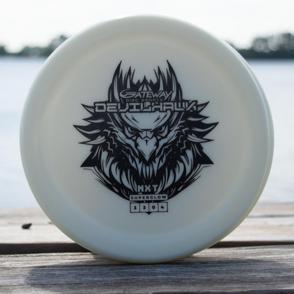 A white Devilhawk - NXT Superglow putter with a black eagle graphic and "Gateway Disc Sports DEJUHawk" stands on wood near water, made from durable Superglow plastic for reliable overstable putt and approach shots.