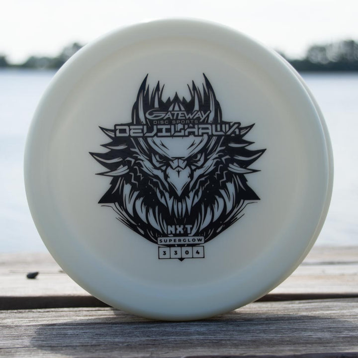 A white Devilhawk - NXT Superglow putter with a black eagle graphic and "Gateway Disc Sports DEJUHawk" stands on wood near water, made from durable Superglow plastic for reliable overstable putt and approach shots.