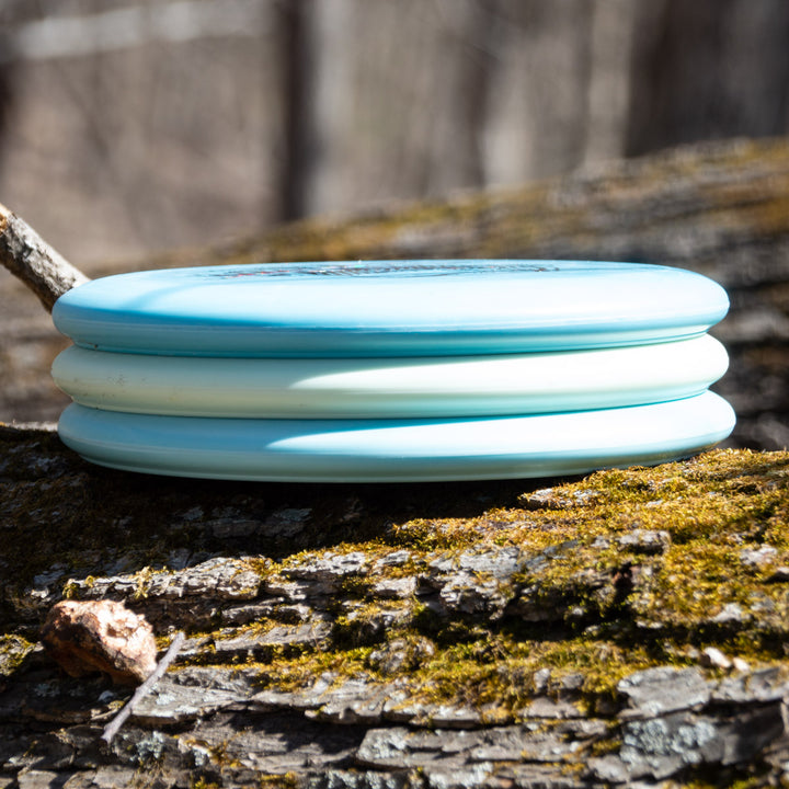 Three light blue Danny Beauchamp's Signature Retro Wizard - SS - Superglow discs are stacked on a mossy tree trunk in a wooded outdoor setting.