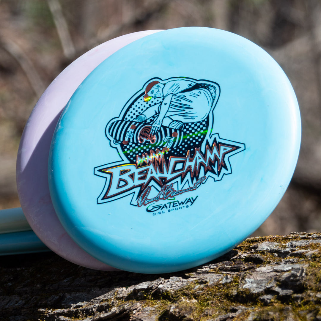 A light blue Danny Beauchamp's Signature Retro Wizard - SS - Superglow disc golf putter with colorful artwork and Gateway Disc Sports branding rests on a tree log.