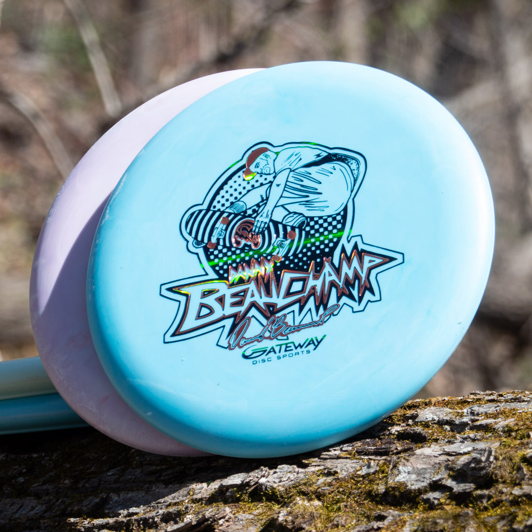 A light blue Danny Beauchamp's Signature Retro Wizard - SS - Superglow disc with a colorful "Bean Champ Gateway Disc Sports" graphic rests on a tree trunk outdoors.