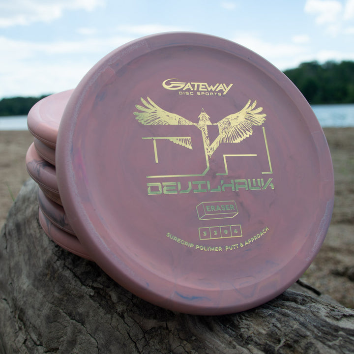 A stack of pink Devilhawk - SureGrip™ Eraser disc golf putters with gold logos sits on a lakeside log, surrounded by trees and blue sky. Enjoy excellent grip and confident putting with the SureGrip™ Eraser blend.