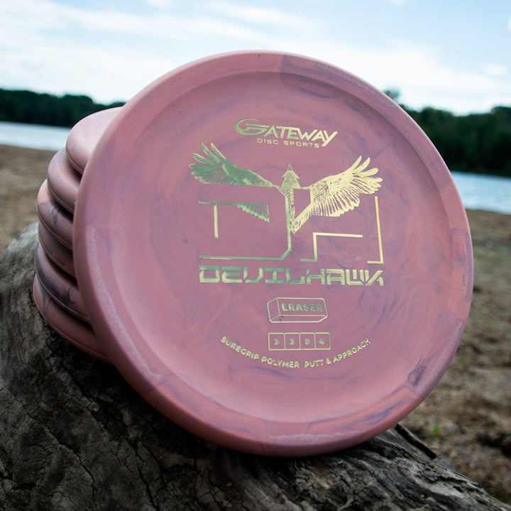 Stack of pink marbled Devilhawk - SureGrip™ Eraser discs with a gold eagle logo, offering superior grip, displayed on a log by a lake under cloudy skies.