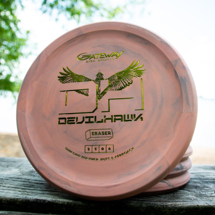 A pink and black marbled Devilhawk - SureGrip™ Eraser disc golf putter, designed for enhanced grip, rests on a wooden surface outdoors near water and trees.