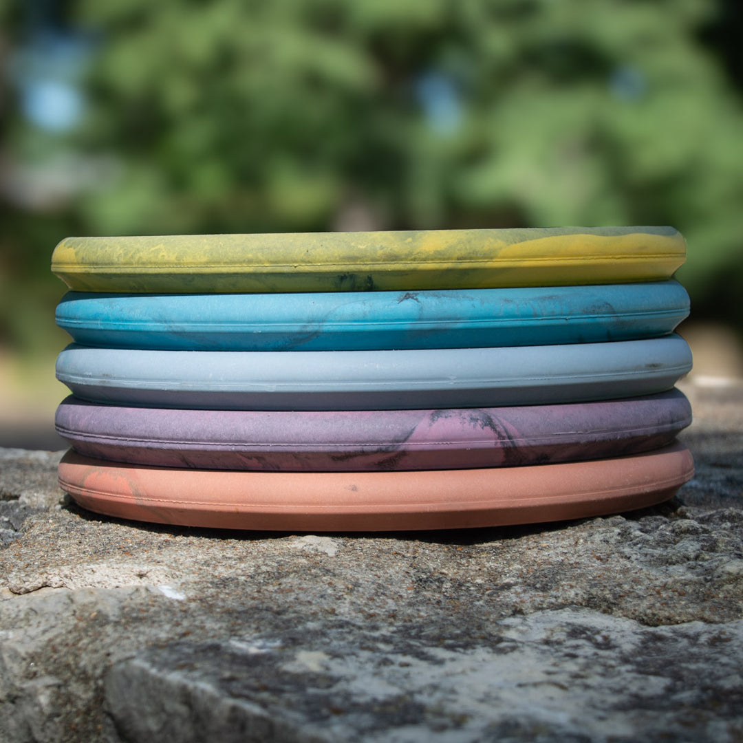 A stack of seven colorful Devilhawk - SureGrip™ Swirly Special Edition frisbees rests on a stone surface, with blurred greenery in the background.