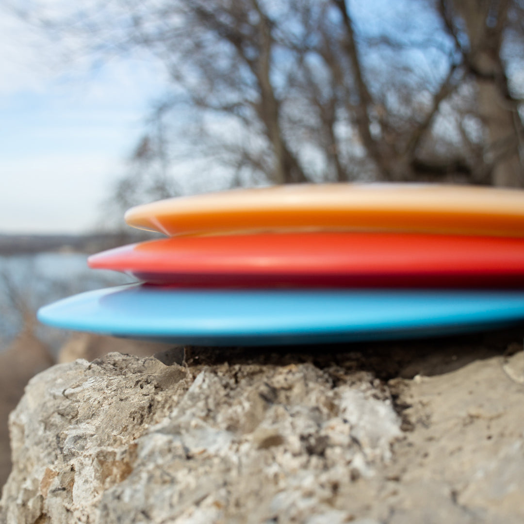 Three Diablo - NXT discs in orange, red, and blue are stacked on a rock with bare trees behind them. Each overstable fairway driver offers control and reliability—perfect for distance flex shots and enhancing your disc golf game.