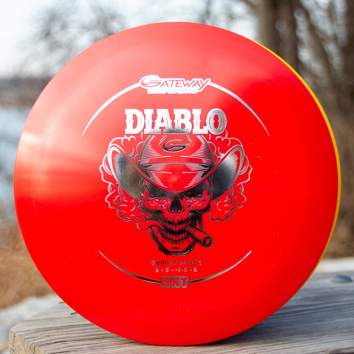 A red Diablo - NXT overstable fairway driver disc golf disc showcases a stylized skull in a cowboy hat, resting on an outdoor wooden surface. Ideal for distance flex shots with dependable control.