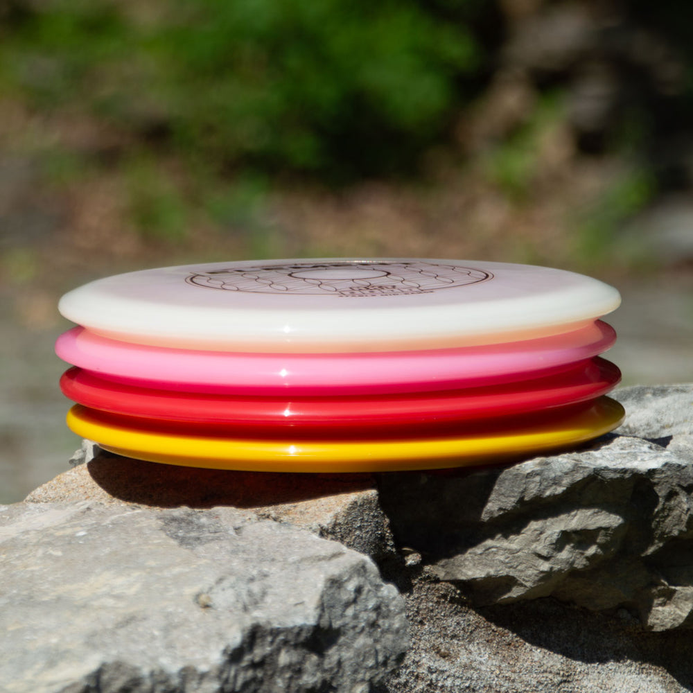 A stack of four Element - NXT discs in white, pink, red, and yellow sits on a rock outdoors amid lush greenery—ideal mid-range discs for players wanting incredible glide and straight flight.