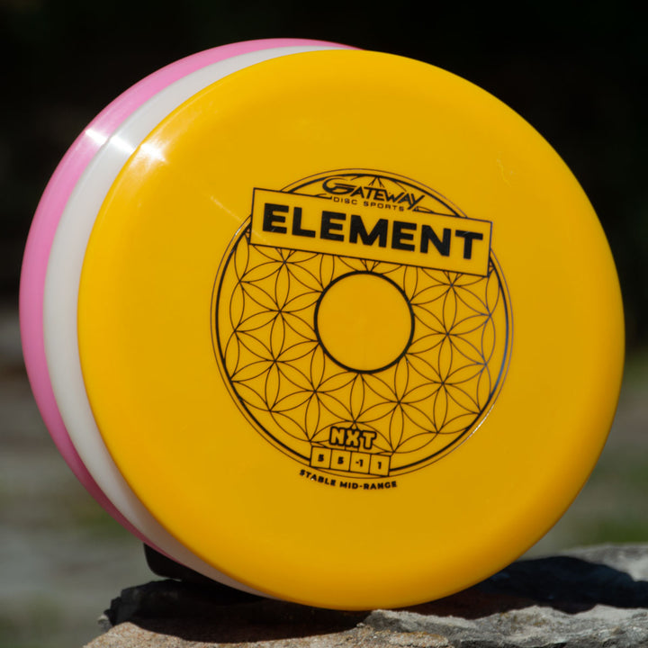 A yellow Gateway Element - NXT mid-range disc with a geometric pattern and logo, stacked on pink and white discs outdoors—engineered for straight flight and incredible glide.