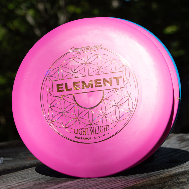 A pink Element Lightweight - SureGrip™  mid-range disc by Gateway Disc Sports, known for its unbelievable glide, rests on a wooden surface with trees blurred in the background.