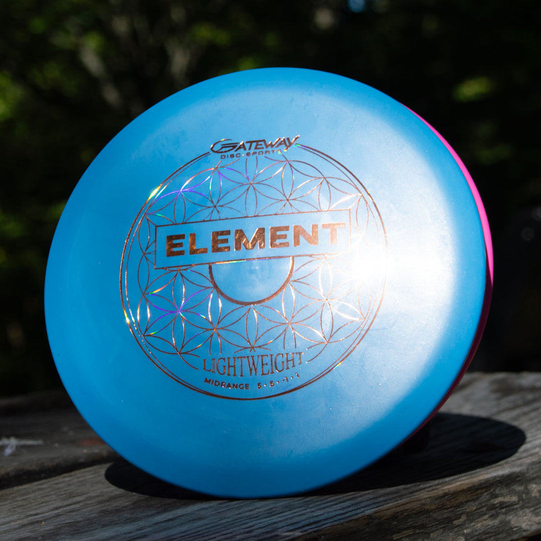 A blue Gateway Disc Sports Element Lightweight - SureGrip™  mid-range disc sits on a wooden surface, sunlight highlighting its design—ideal for players wanting incredible glide and straight flight.