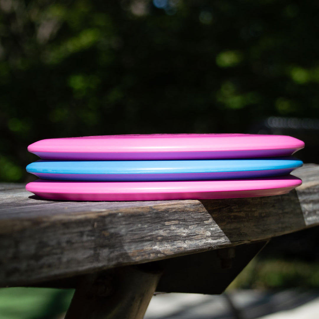The Element Lightweight - SureGrip™  mid-range disc, celebrated for its straight flight and impressive glide, is stacked with two other plastic flying discs on a wooden bench outside, greenery visible in the background.