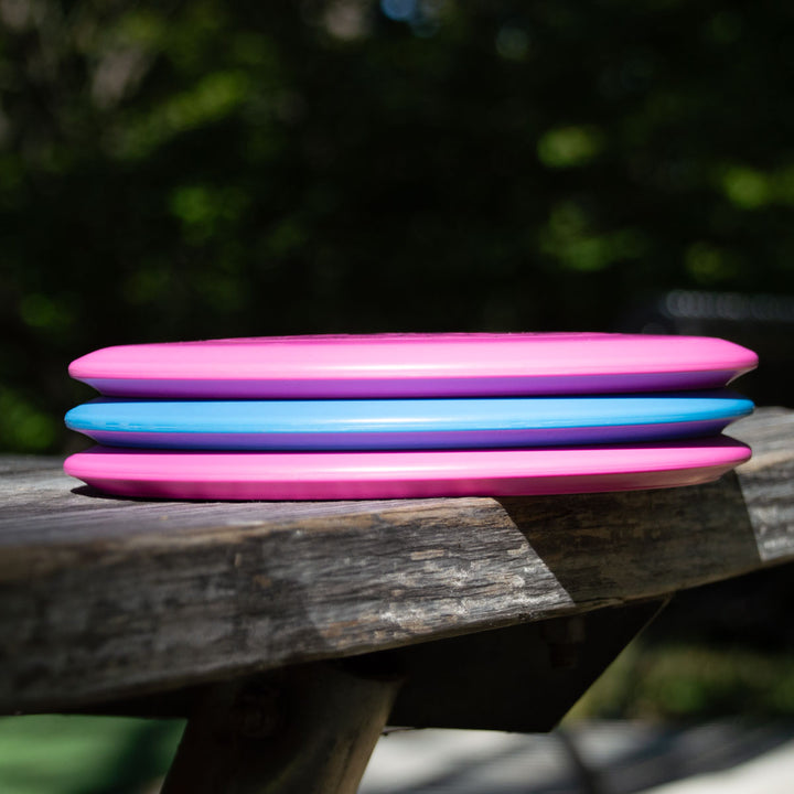 The Element Lightweight - SureGrip™  mid-range disc, celebrated for its straight flight and impressive glide, is stacked with two other plastic flying discs on a wooden bench outside, greenery visible in the background.