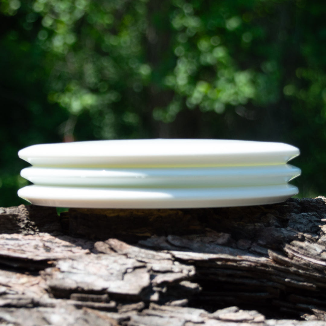 Three white Dye Canvas Element - NXT ceramic plates are stacked on a rough tree trunk outdoors, with a blurred green leafy background.