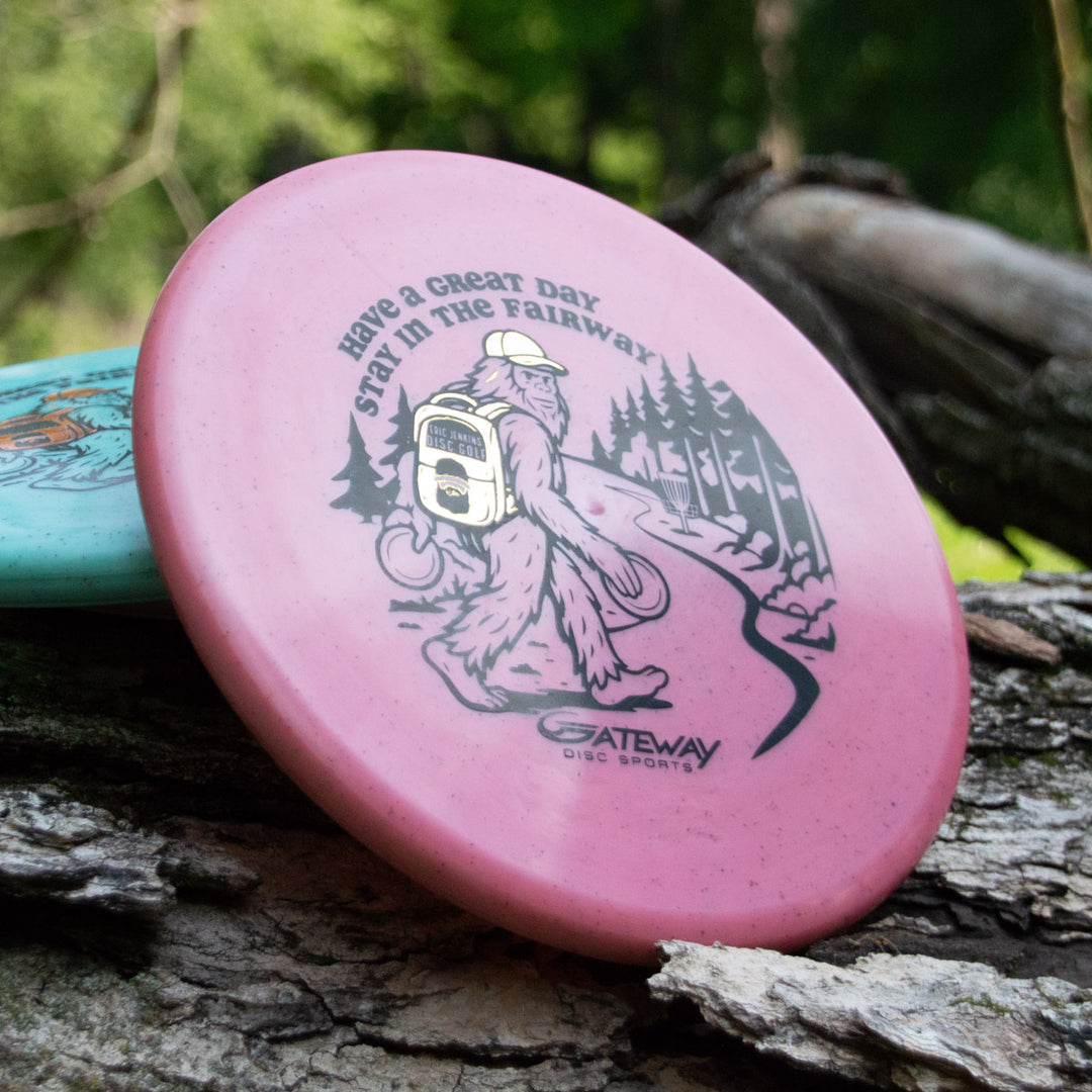 A pink Eric Jenkins Signature Series Chief - Diamond Hemp low profile putter disc featuring Bigfoot and the phrase "Have a Great Day, Stay in the Fairway" rests on a tree trunk.