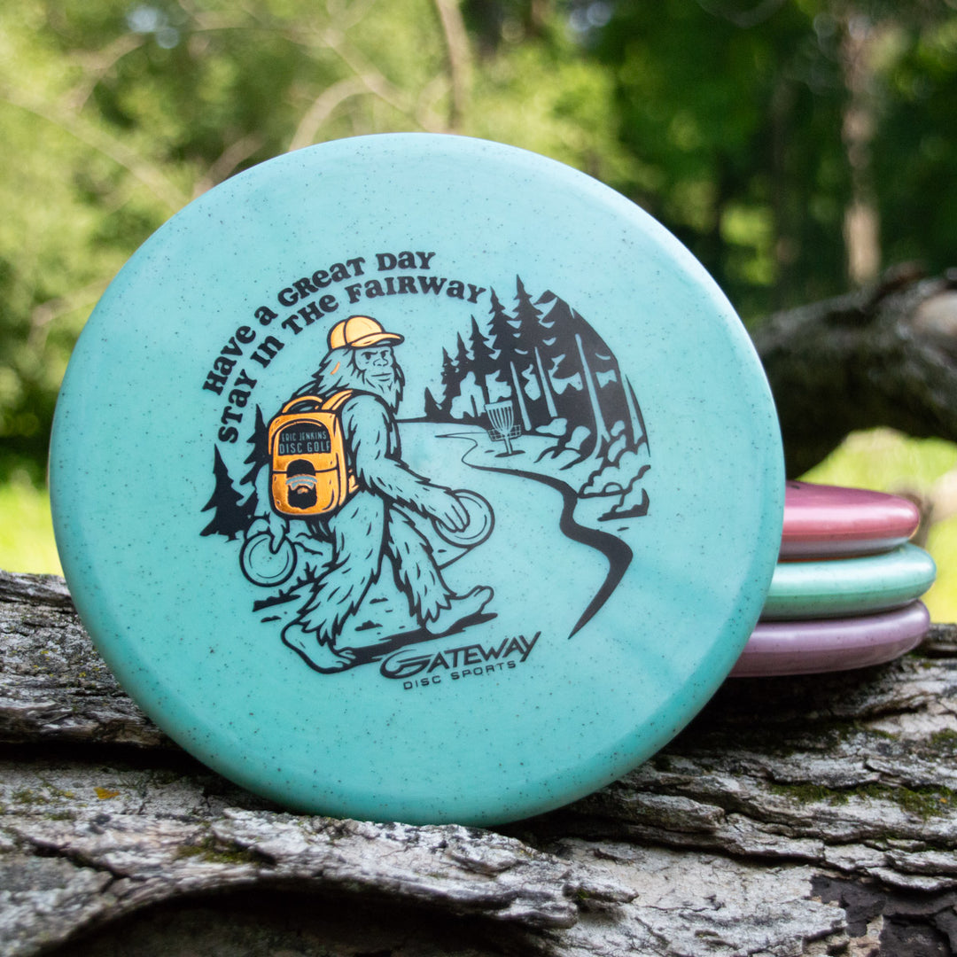 A turquoise Eric Jenkins Signature Series Chief - Diamond Hemp putter shows Bigfoot with a disc bag and the phrase "Have a Great Day Stay in the Fairway," relaxing on a log outdoors.